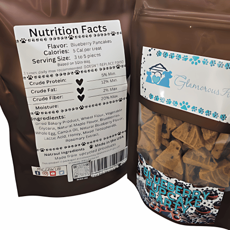 Close-up view of the nutrition facts label on a bag of Blueberry Pupcake Party by Glamorous Pups, detailing ingredients like dried bakery product, wheat flour, and blueberry flavor. The package also shows a serving suggestion of 3 to 5 pancakes for a 50lb dog, emphasizing the product's low calorie and high crude protein content. The packaging design includes the Glamorous Pups logo and a visible window displaying the treats, highlighting the all-natural ingredients and made-in-the-USA quality assurance.