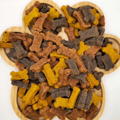 Feast of Beasts Pup-Pleaser Dog Treats by Glamorous Pups artfully displayed on a bamboo serving board. This mixture showcases various shaped treats in shades of gold, brown, and gray, all made with high-quality beef, turkey, and chicken, perfect for rewarding your furry friend with a savory snack.