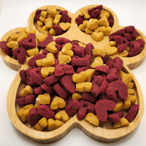 Assorted Pup's Love Kisses Dog Treats by Glamorous Pups, displayed on a heart-shaped wooden serving board, featuring vibrant red and yellow treats crafted for pet nutrition and satisfaction.