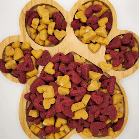 Colorful display of Pup's Love Kisses Dog Treats by Glamorous Pups arranged on a heart-shaped bamboo board, featuring delicious red and yellow heart-shaped treats tailored for canine enjoyment.