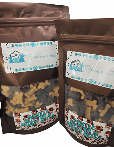 Two packages of Glamorous Pups branded dog treats displayed prominently. The bags, in a glossy brown finish, showcase visible, healthy dog biscuits in shapes of bones and paws through a clear window. The labels feature the Glamorous Pups logo and the product name 'Gentle Berry Bites,' emphasizing natural and gentle ingredients suitable for all dogs.