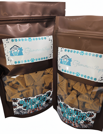 Two bags of Blueberry Pupcake Party by Glamorous Pups in 1lb and 8oz sizes. Each glossy brown package has a clear window showcasing blueberry-infused, bone-shaped dog treats. Labels feature the Glamorous Pups logo and blue paw prints, emphasizing all-natural ingredients and delicious blueberry flavor, ideal for pet owners who value health and taste in pet snacks.