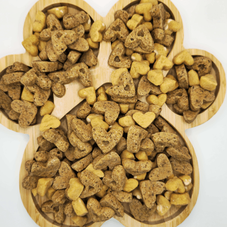 Sweet À La Mode Dog Treats by Glamorous Pups arranged on a wooden paw print-shaped serving board. These treats, in heart and bone shapes, vary in color from golden to dark brown, highlighting their natural ingredients and appealing texture. Ideal for pet owners seeking gourmet, nutritious snacks, these treats emphasize the creative and high-quality standards of Glamorous Pups, made with all-natural ingredients in the USA.