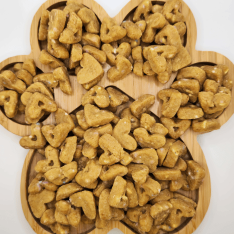 Assorted Pup Cake Confetti Cravings Dog Treats by Glamorous Pups displayed on a wooden paw print-shaped serving board. The treats, shaped like small birthday cakes, feature a golden texture with visible confetti bits, highlighting their fun and festive nature. Perfect for pet owners seeking unique, high-quality treats made in the USA, these snacks emphasize Glamorous Pups’ commitment to gourmet, all-natural ingredients.