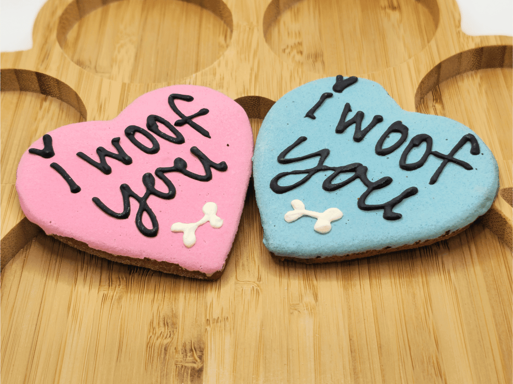 Presented on a wooden paw-shaped serving board, these 'Cherished Woof Hearts' by Glamorous Pups include one pink and one blue specialty dog treat. Each heart-shaped biscuit is beautifully iced with 'I woof you' in elegant black lettering, accented with adorable white bone details, perfect for showing love to your furry friend.