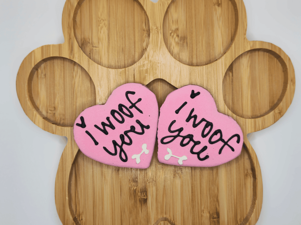 Delightful image of two pink, heart-shaped specialty dog treats named 'Cherished Woof Hearts' by Glamorous Pups, placed on a wooden serving board shaped like a paw. Each treat is lovingly decorated with the phrase 'I woof you' in elegant black icing, adorned with cute white bone accents, perfect for showing love to your furry friend.